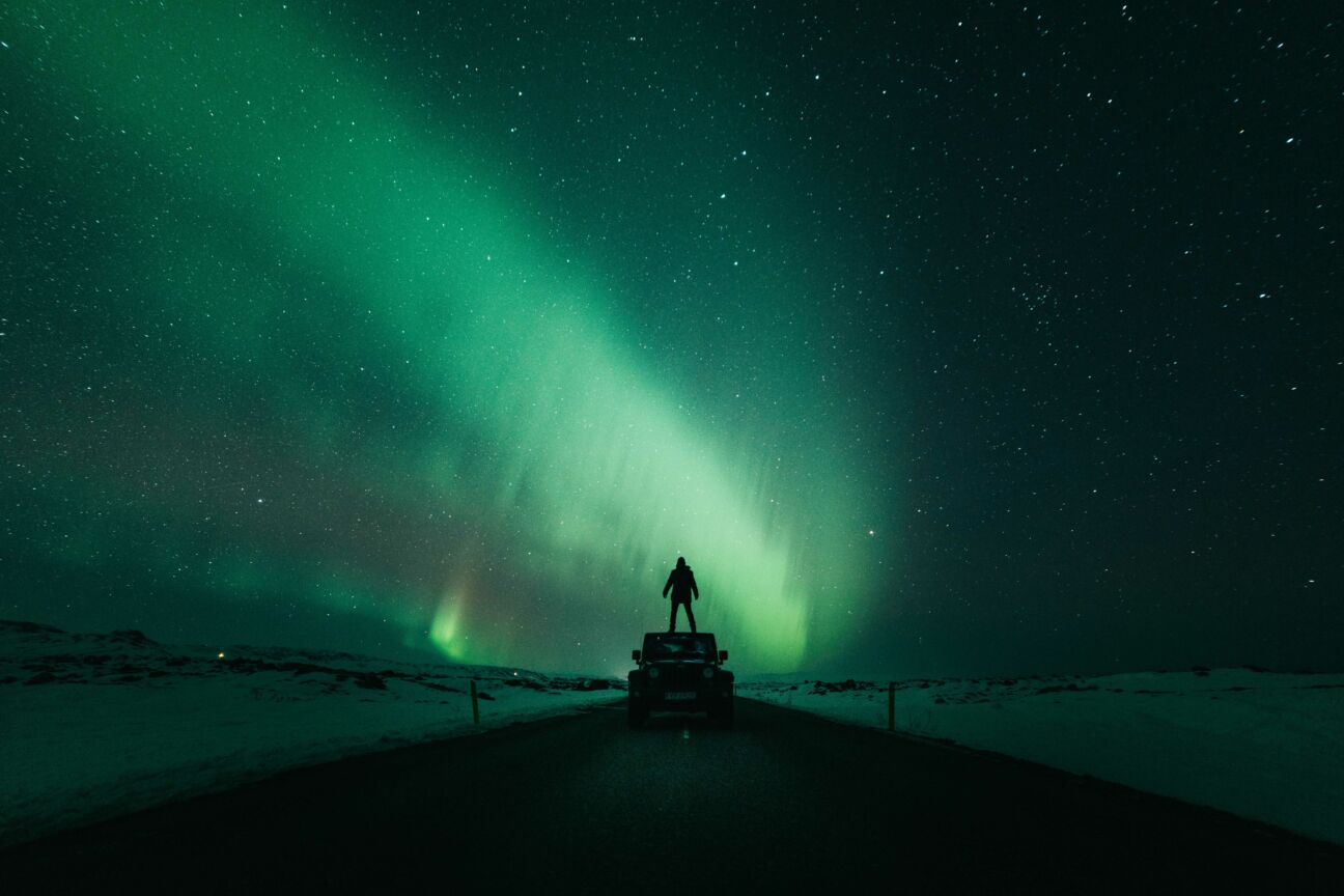 man stands on top of jeep while northern lights and stars are in the night sky above